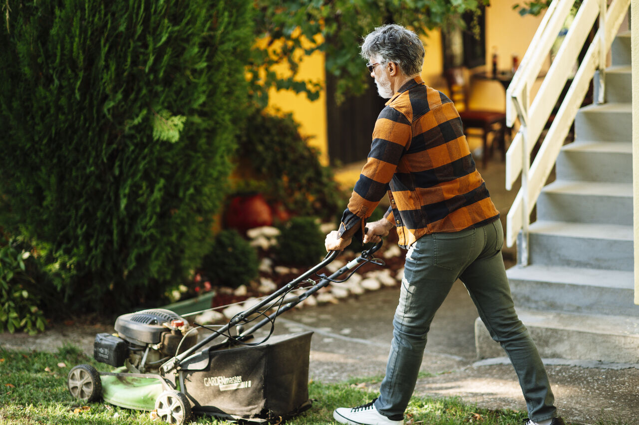Gardener pushing a lawn mower beside garden beds at a suburban home