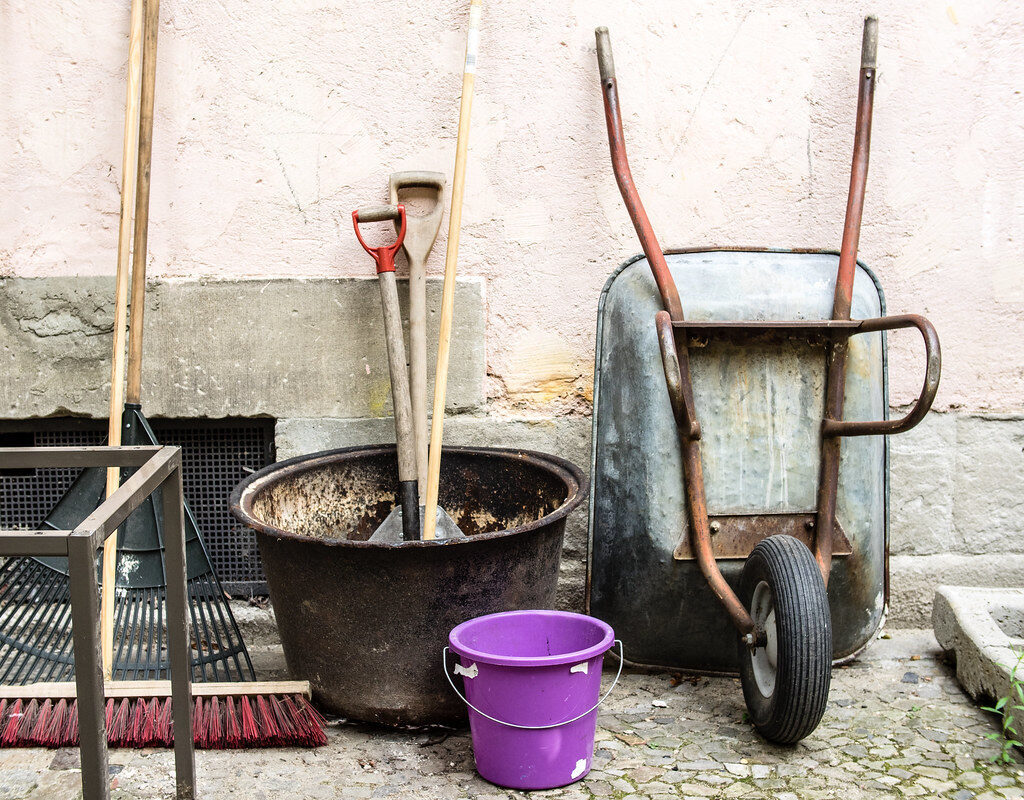 Work-worn spades, rake, broom, bucket and wheelbarrow by a wall